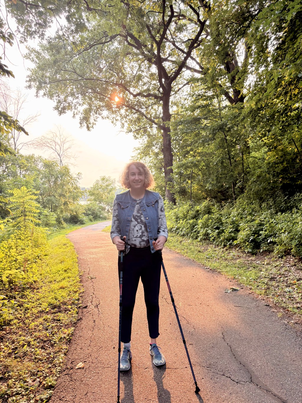 Dielle Lundberg, a white transfeminine person walking with her walking sticks on a trail in the woods